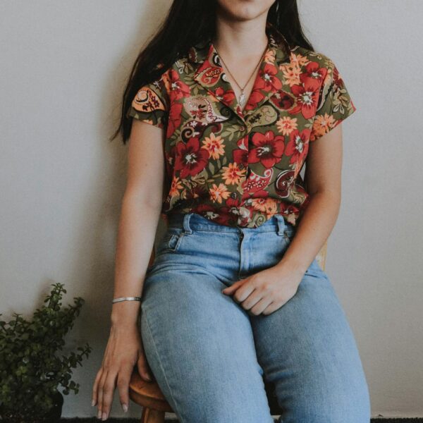 Woman sitting on stool wearing floral shirt and jeans indoors with a plant by her side.
